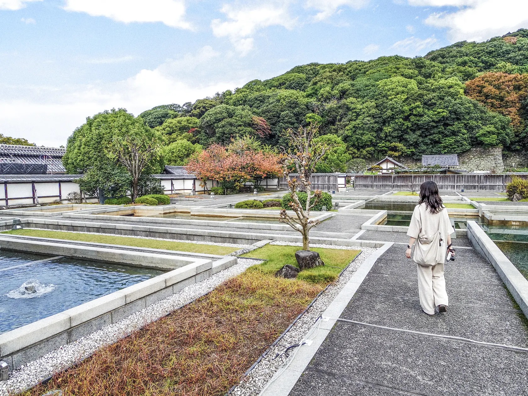 堀之内の庭園。石組みの水面と紅葉しはじめた木々、背後に城山の緑。園路を人物が歩いている。