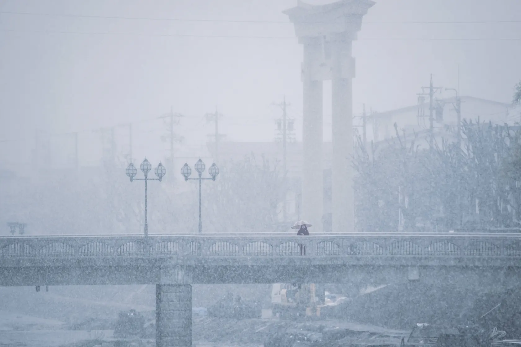 雪の降りしきる高山の中橋。白くかすむ橋の上に、傘の人影がぽつんと見える。