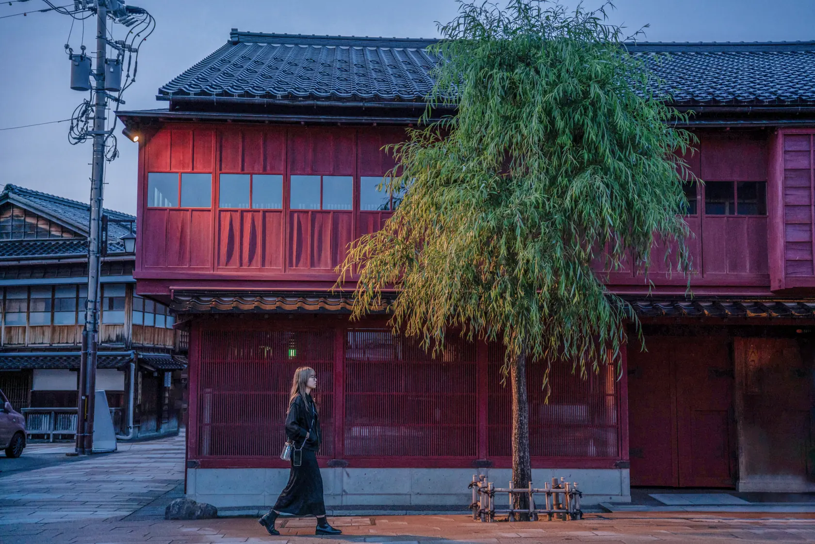 夕暮れの弁柄格子の茶屋建築と柳の木。誰もいない通りを、黒い服の巡礼者が一人歩いている。
