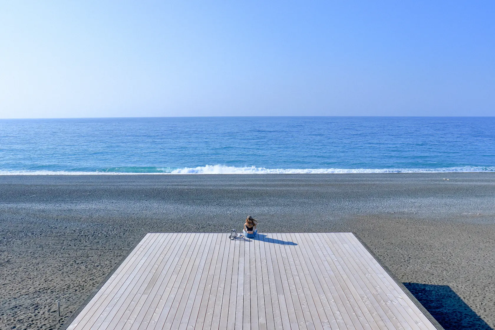 青い海へ突き出た白木のデッキ。その先端に人物が座って海を見ている。