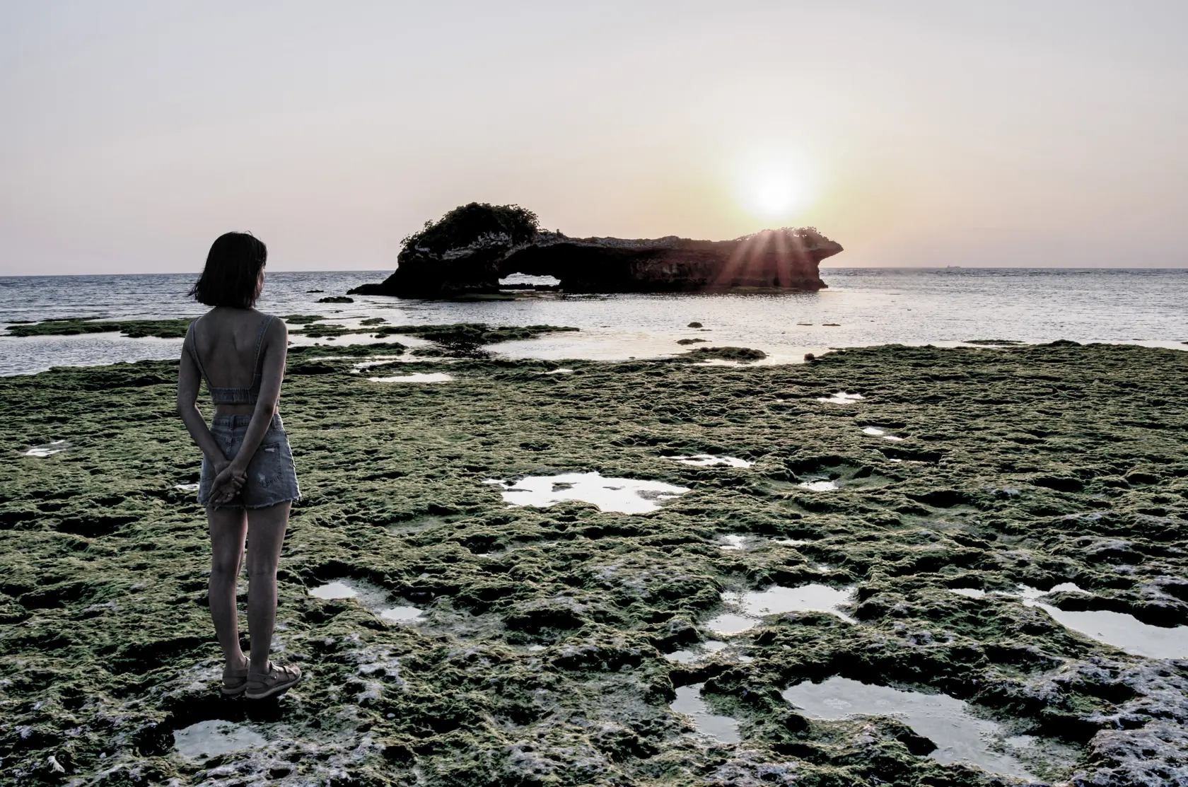 夕日が沈む渡具知の海。岩礁の上に人物が立ち、水平線を眺めている。