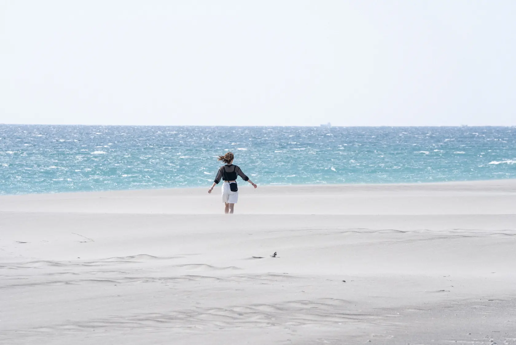 昼の鳥取砂丘。白い砂の向こうの日本海へ、風に髪をなびかせて歩いていく巡礼者の後ろ姿。